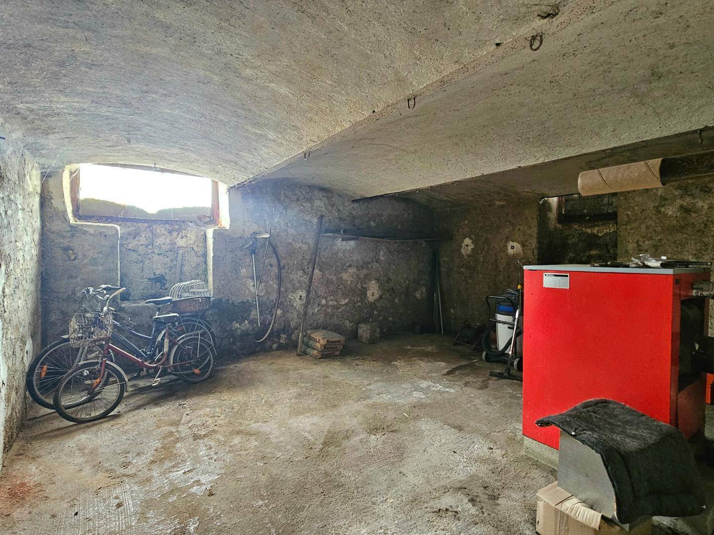 Alter Kellerraum mit Gewölbedecke, Betonboden, Fahrrädern und einem roten Heizkessel.