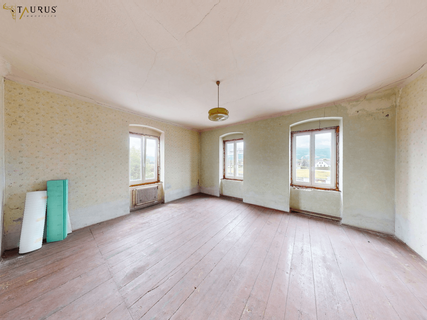 Geräumiges Zimmer mit altem Holzboden und drei Fenstern, bereit für Renovierung.