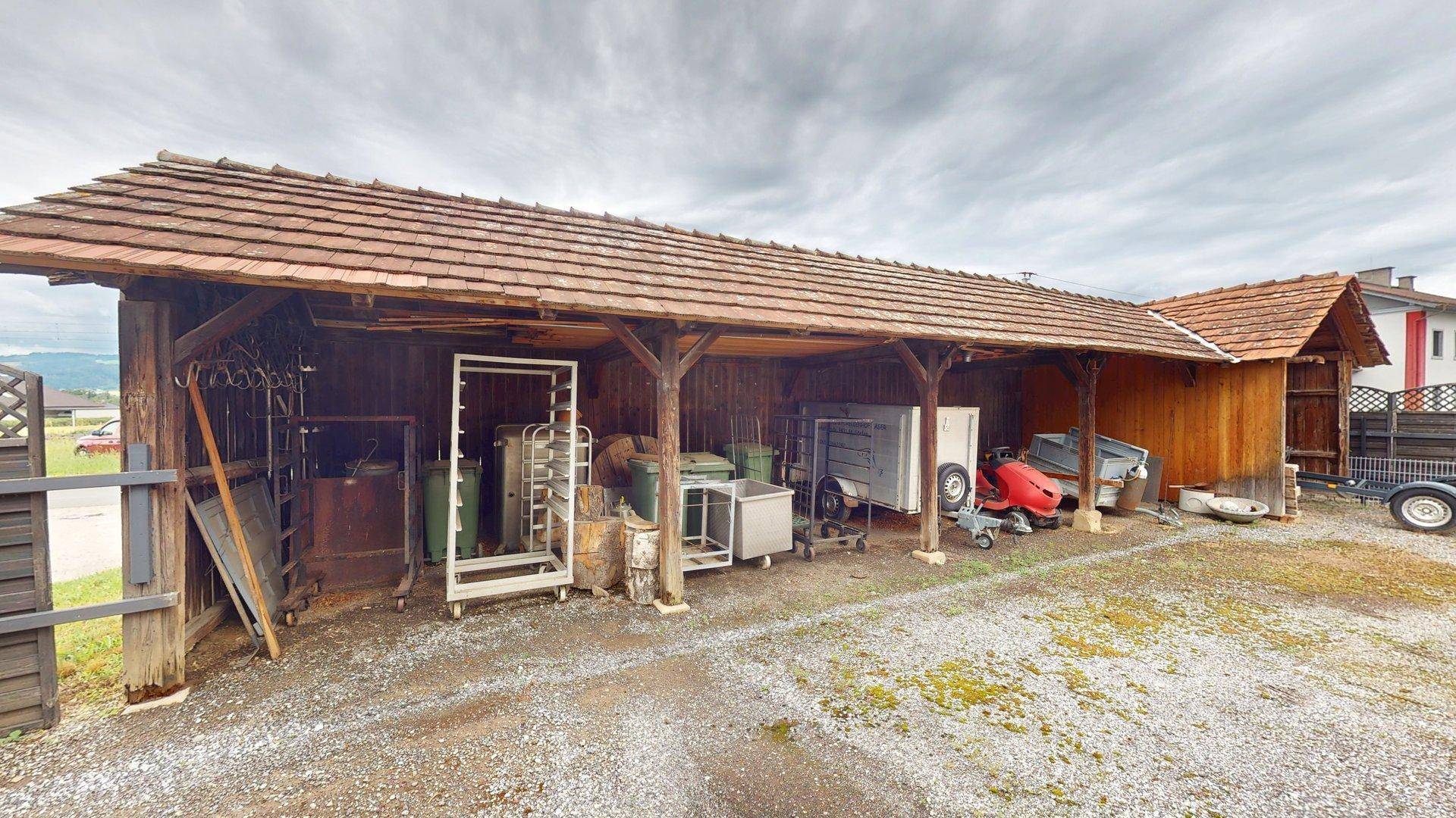 Offener Holzschuppen mit Ziegeldach, genutzt als Unterstand für diverse Geräte und Fahrzeuge.