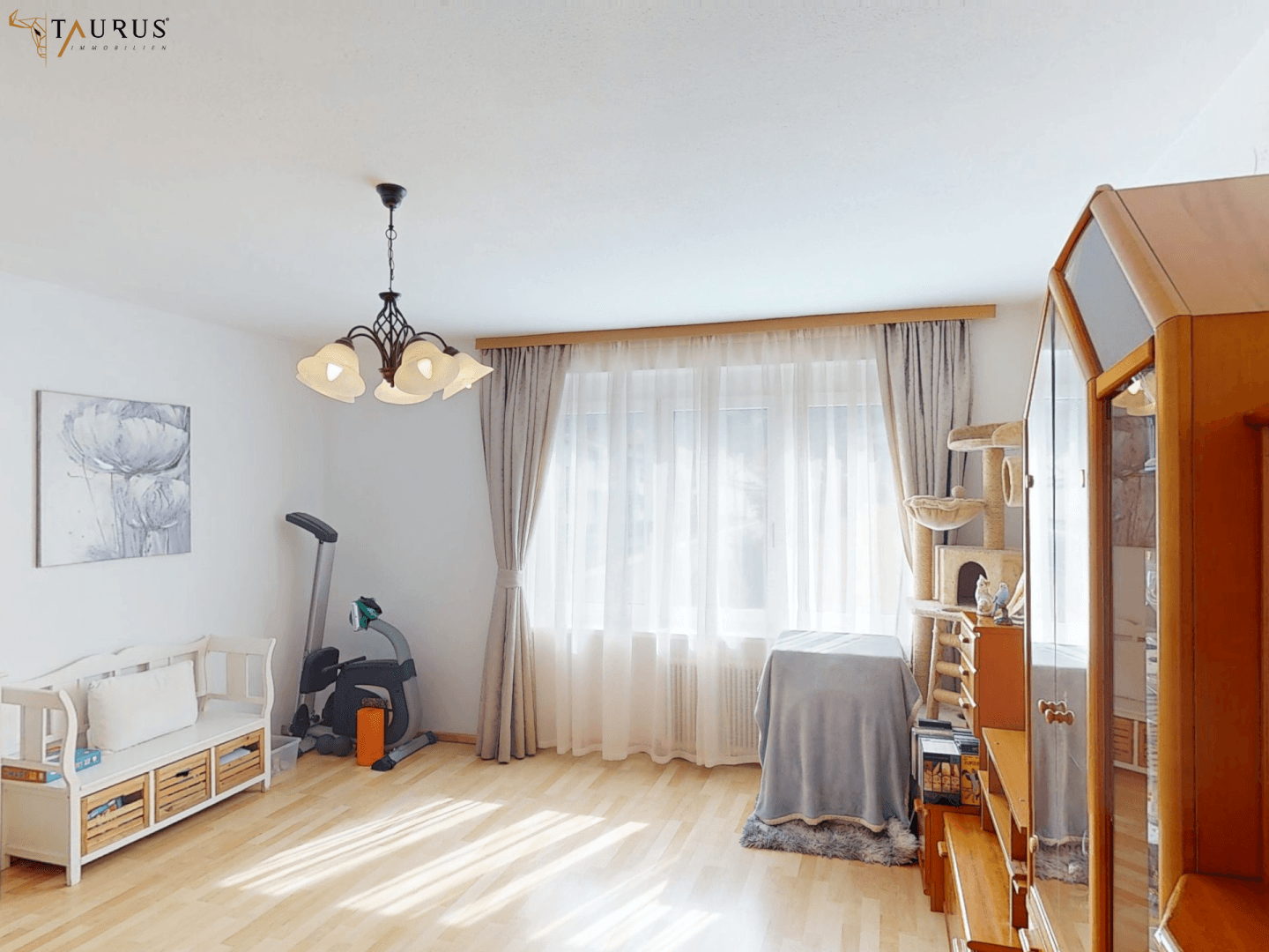 Helles Wohnzimmer mit Holzfußboden, großen Fenstern und einem Fitnessgerät, gemütlich eingerichtet.