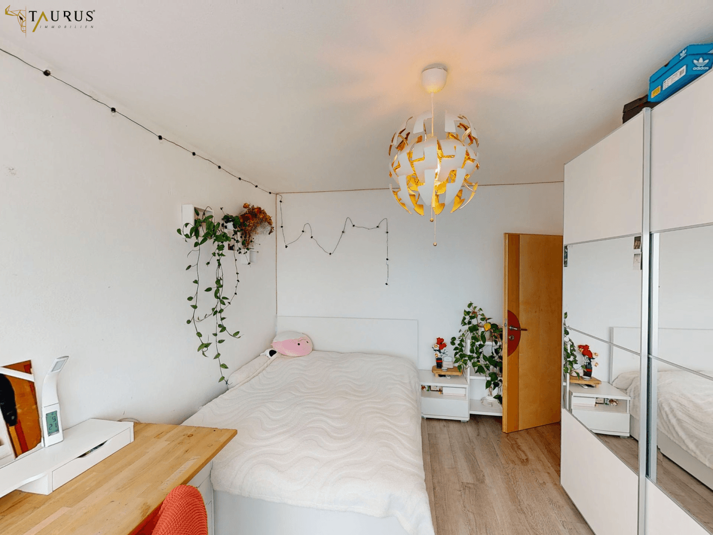 Gemütliches Schlafzimmer mit weißem Bett, Schreibtisch und einem Spiegelschrank, dekoriert mit Pflanzen.