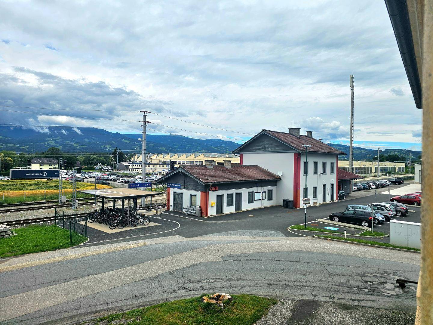 Blick auf einen Bahnhof mit Gleisen, Parkplätzen und umliegenden Gebäuden, im Hintergrund Berge.