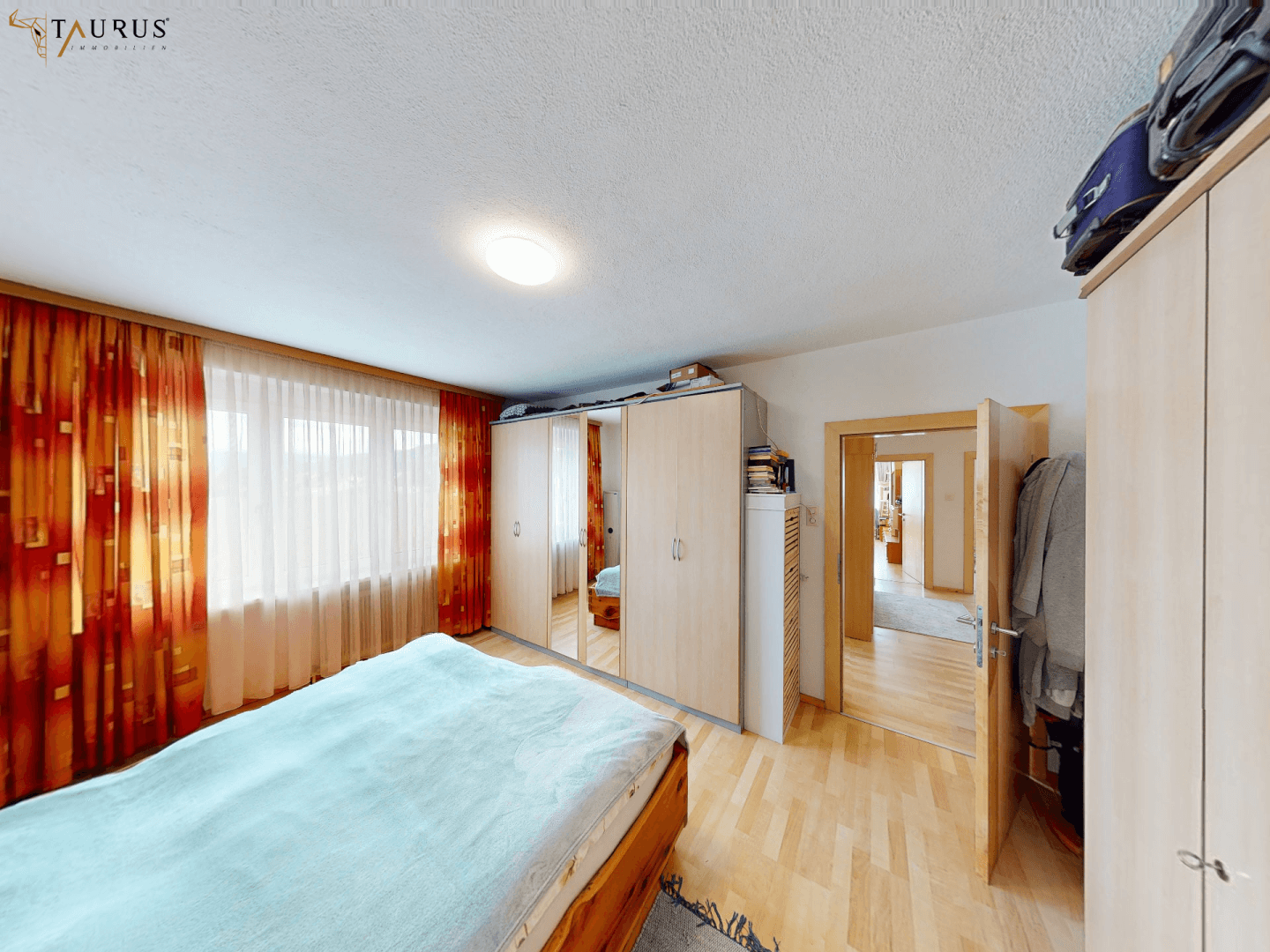 Geräumiges Schlafzimmer mit Doppelbett, Holzschrank und großen Fenstern mit orangefarbenen Vorhängen.