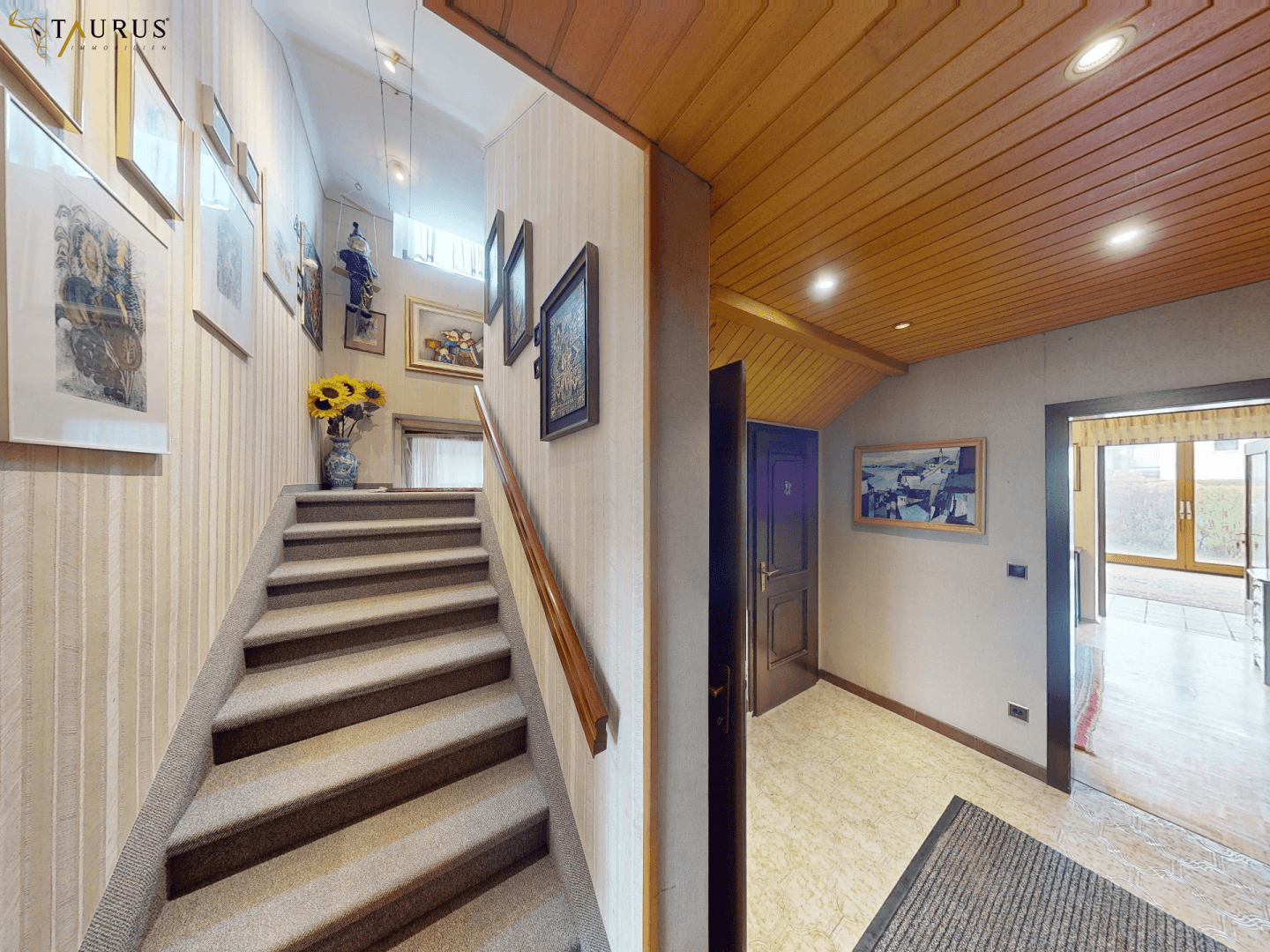 Treppenhaus mit Teppichboden, Holzgeländer und Bildern an den Wänden.