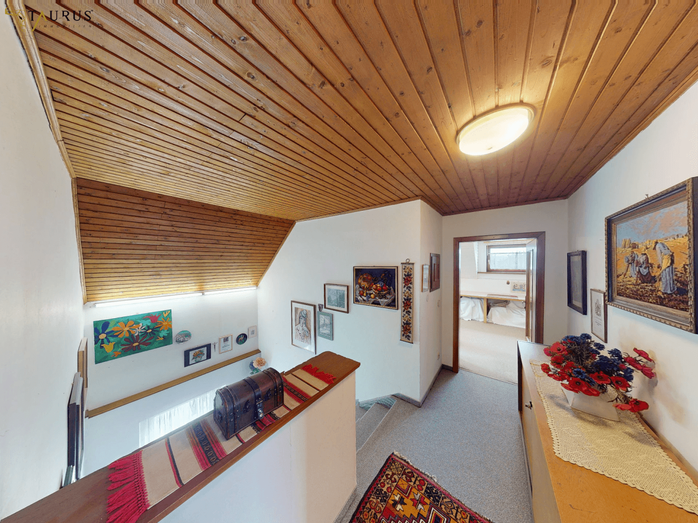 Heller Treppenaufgang mit Holzpaneeldecke und dekorativen Bildern an den Wänden.