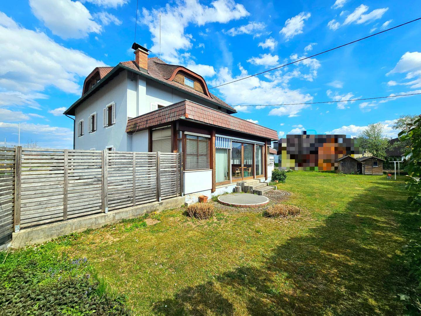 Seitenansicht des Hauses mit Garten, Holzzaun und sonnigem Rasen unter blauem Himmel.