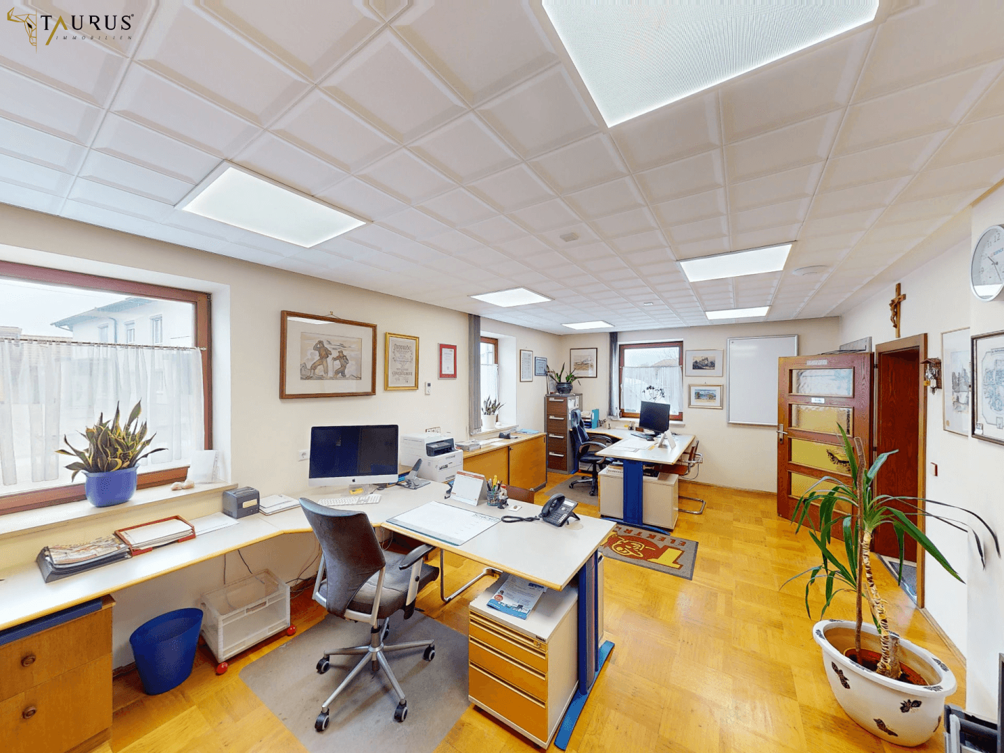 Helles Büro mit mehreren Arbeitsplätzen, Holzparkett und großen Fenstern.