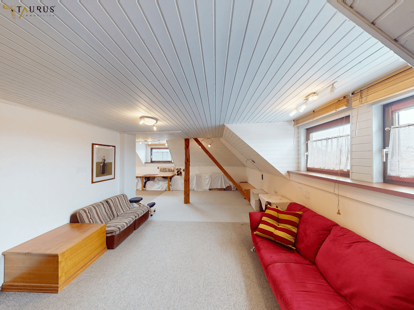 Geräumiges Dachgeschoss-Wohnzimmer mit Teppichboden, weißen Holzdecken und mehreren Fenstern.