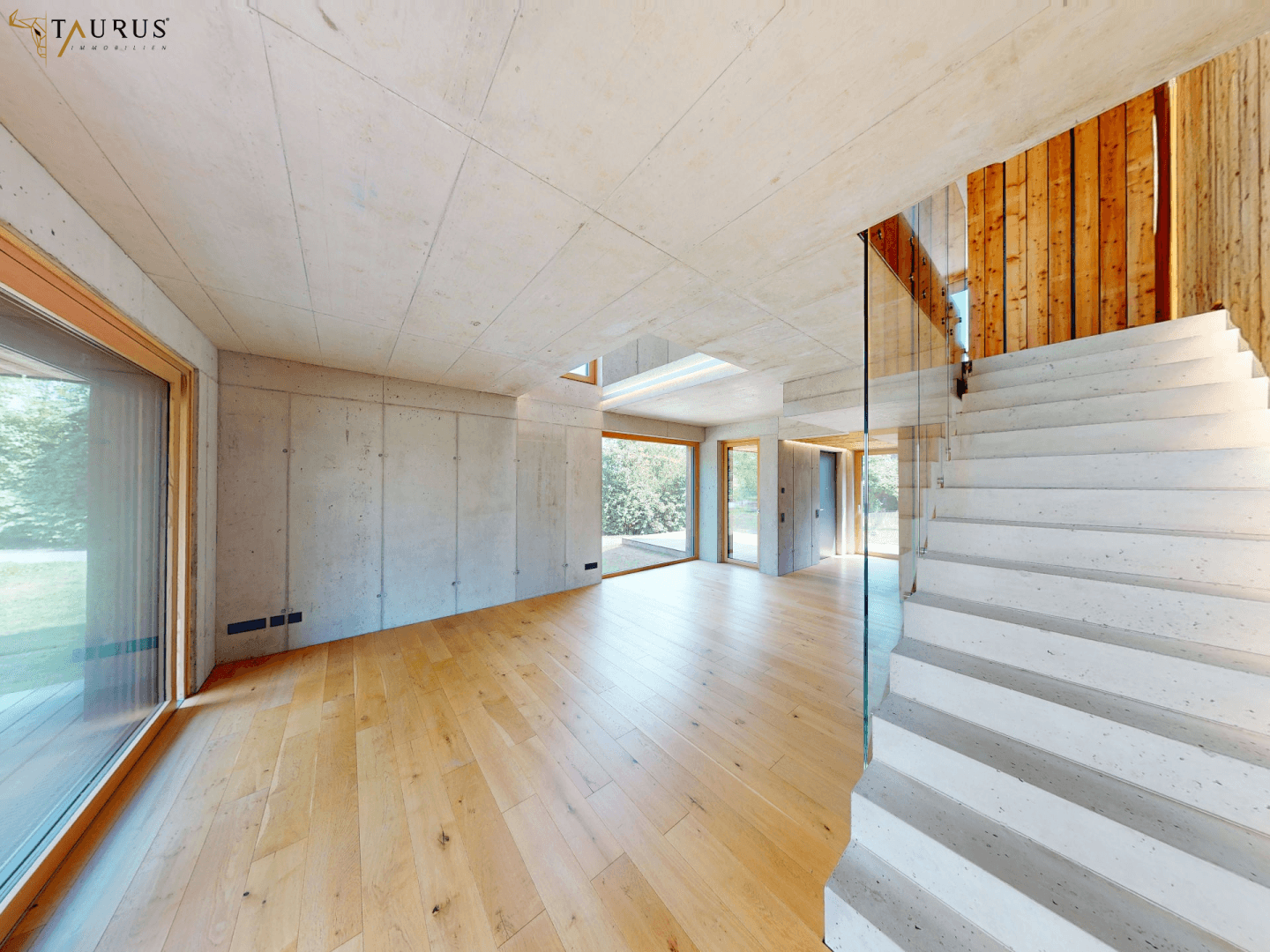 Weitläufiger Wohnbereich mit Holzboden, Sichtbetonwänden und moderner Treppe.