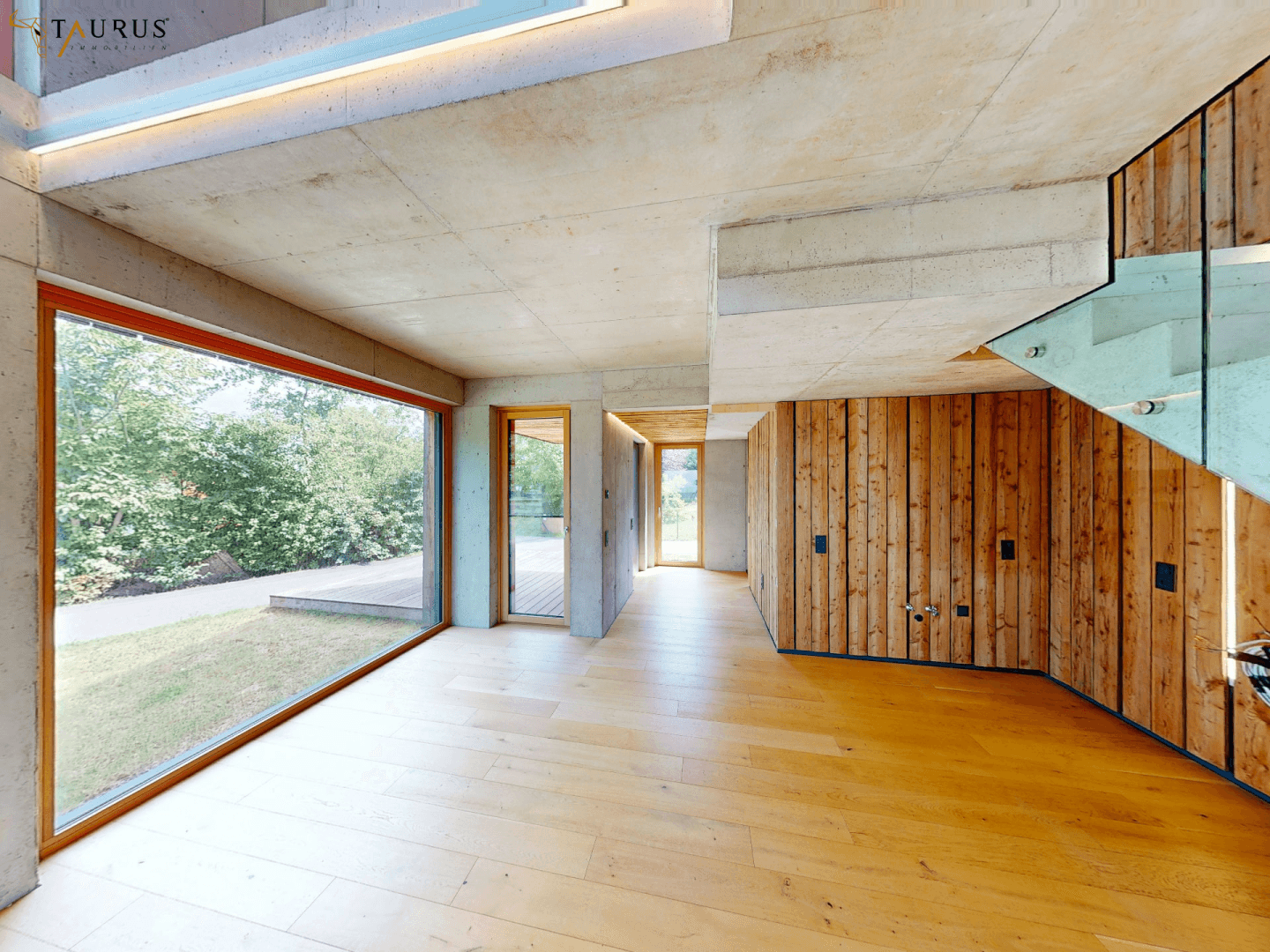 Offener Wohnbereich mit Holzboden, Sichtbetonwänden und Zugang zum Garten durch große Fenster.