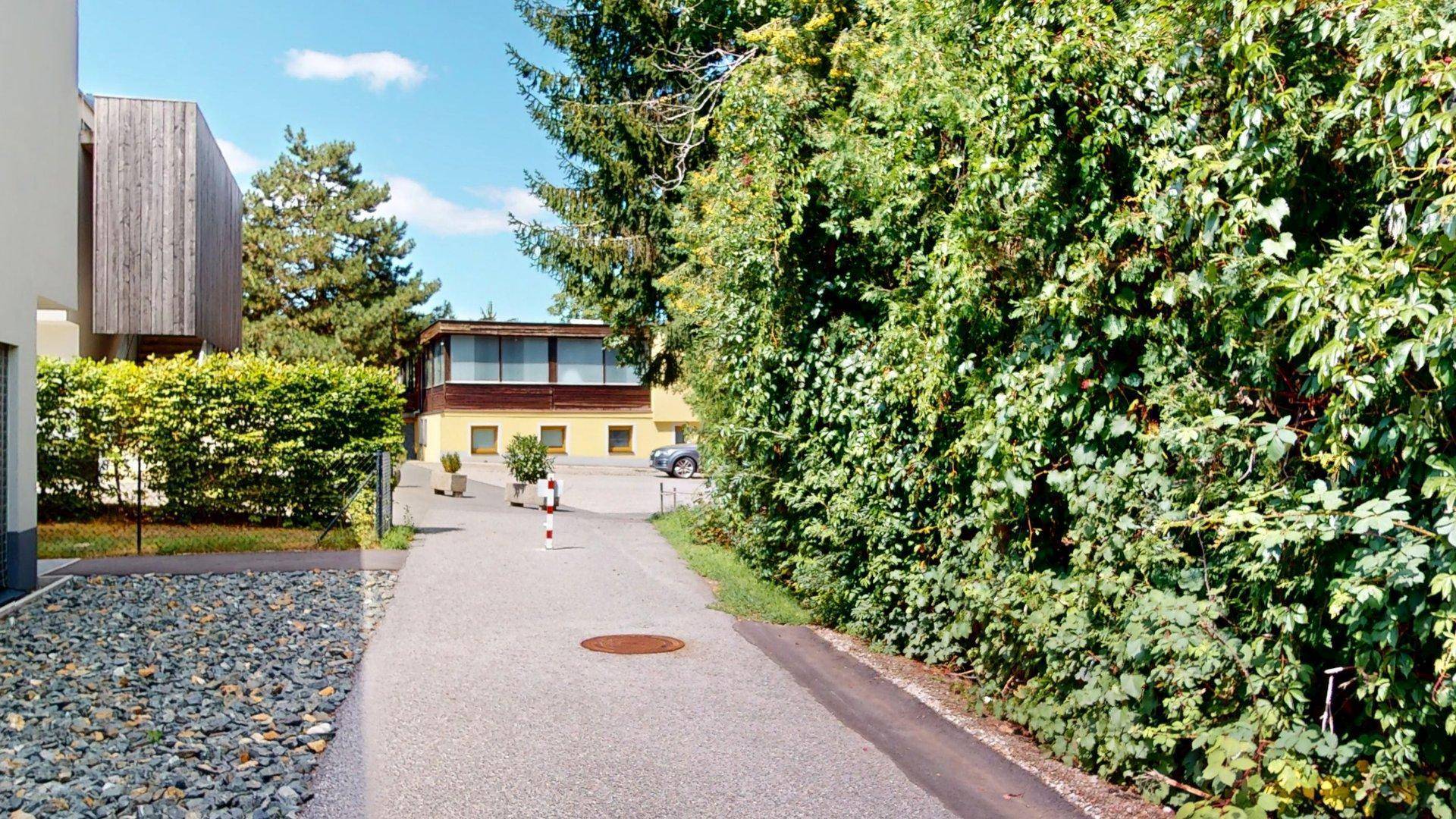 Zufahrtsweg zu einem Wohngebiet mit viel Grün und einem weiteren Gebäude im Hintergrund.