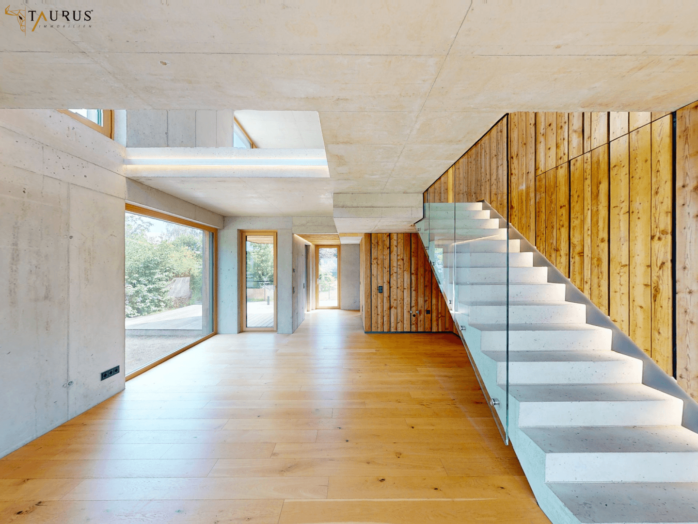 Geräumiger Wohnbereich mit Holzboden, Sichtbetonwänden und moderner Treppe mit Glasgeländer.