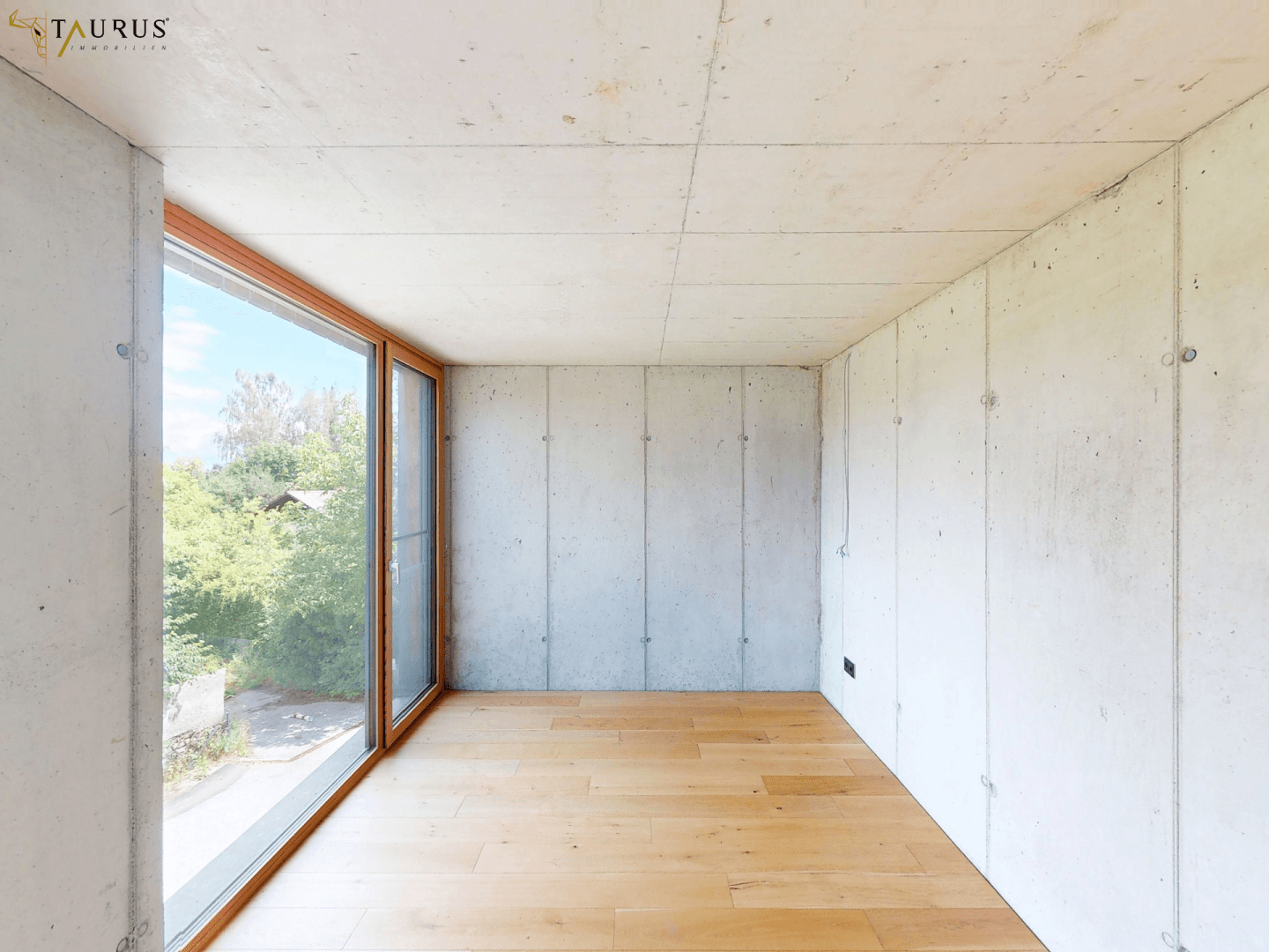 Leeres Zimmer mit Holzboden, Sichtbetonwänden und großen Fenstern mit Blick ins Grüne.