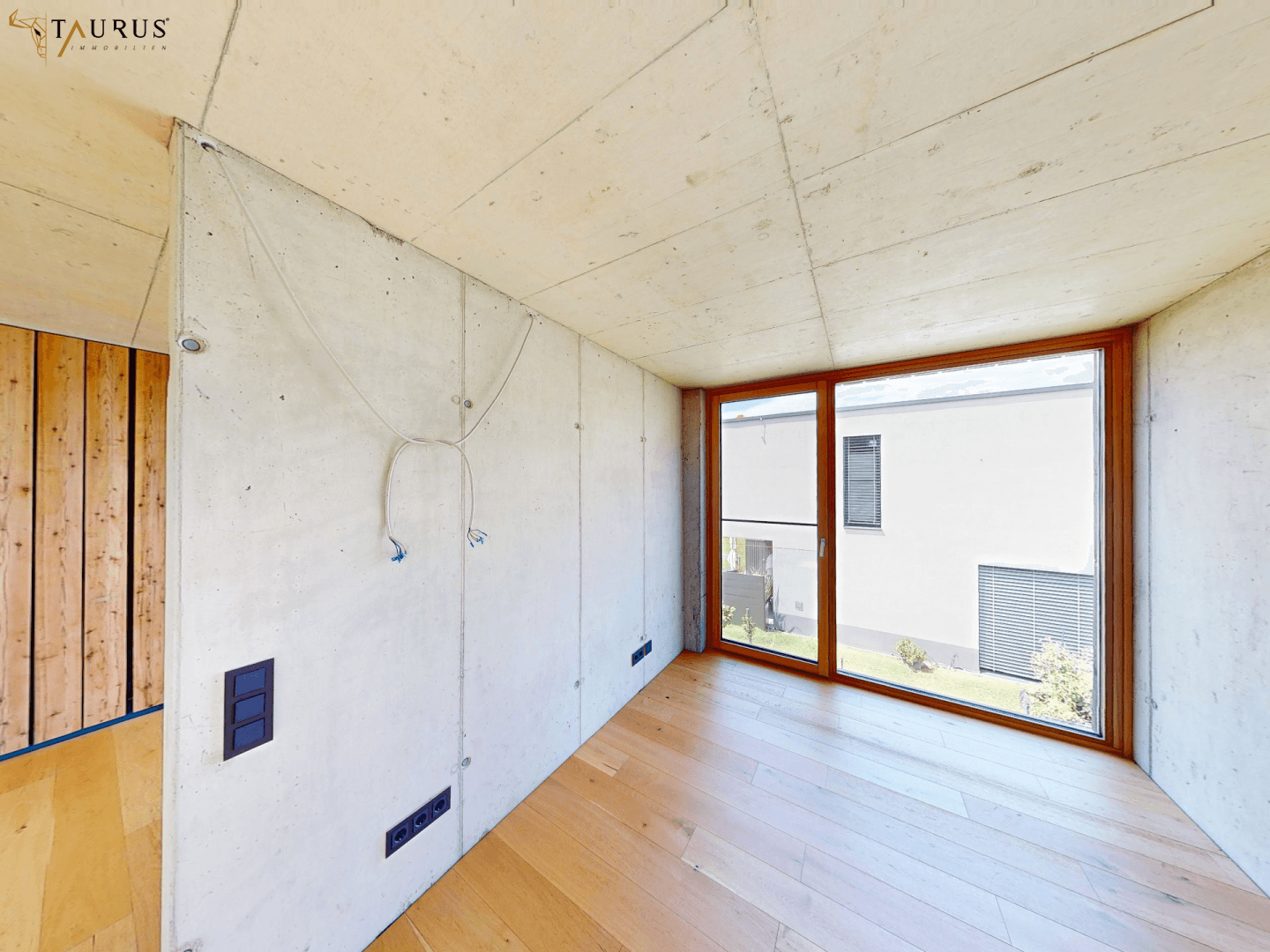 Leeres Zimmer mit Holzboden, Sichtbetonwänden und großen Fenstern mit Blick auf die Nachbarschaft.