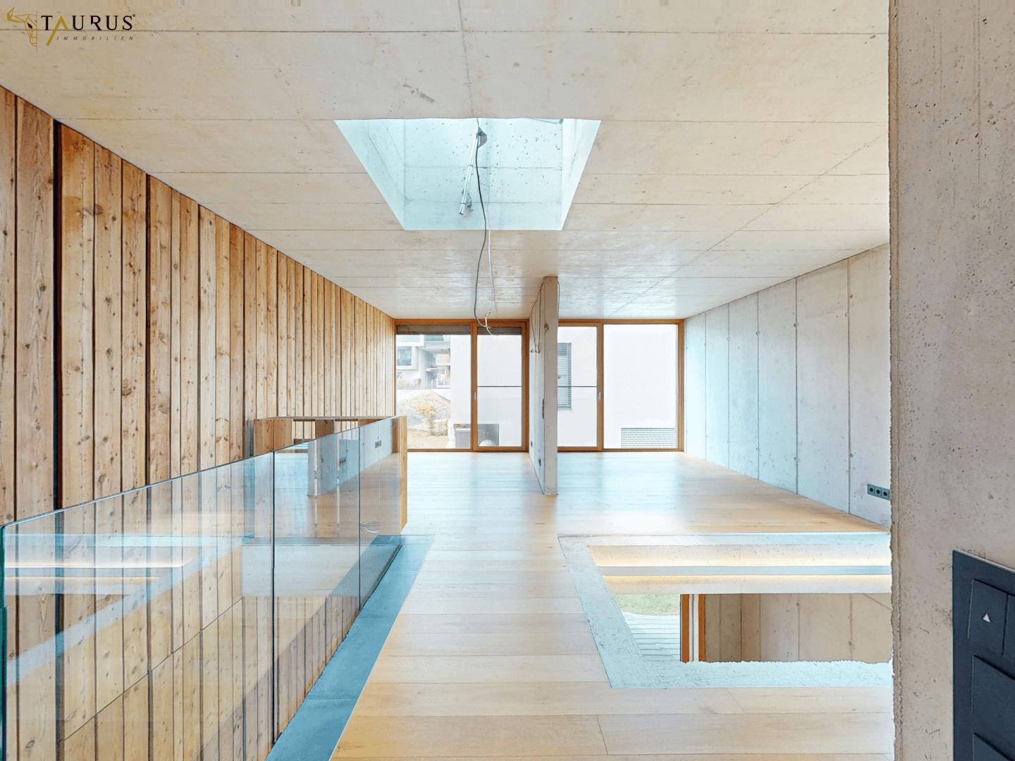 Heller Flur im Obergeschoss mit Holzboden, Sichtbeton und Glasgeländer.