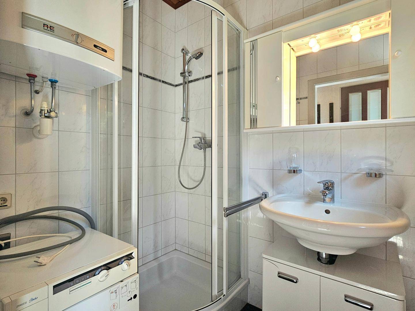 Kleines Badezimmer mit Duschkabine, Waschbecken und Waschmaschine, hell gefliest.