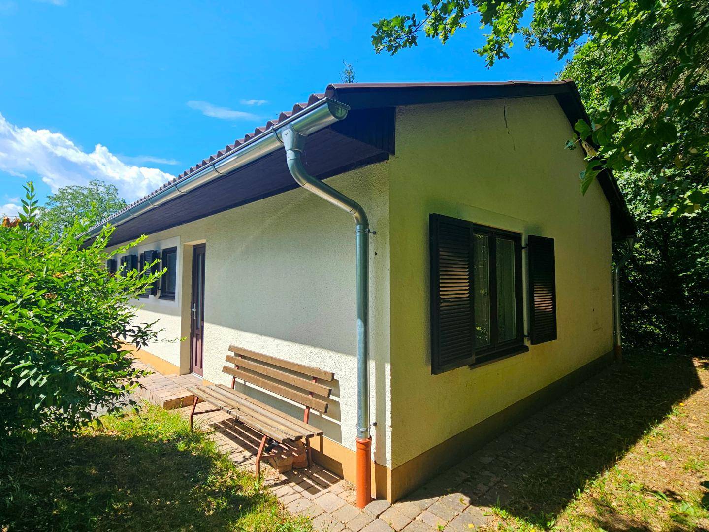 Seitenansicht des Hauses mit Regenrinne, Holzbank und grüner Umgebung.