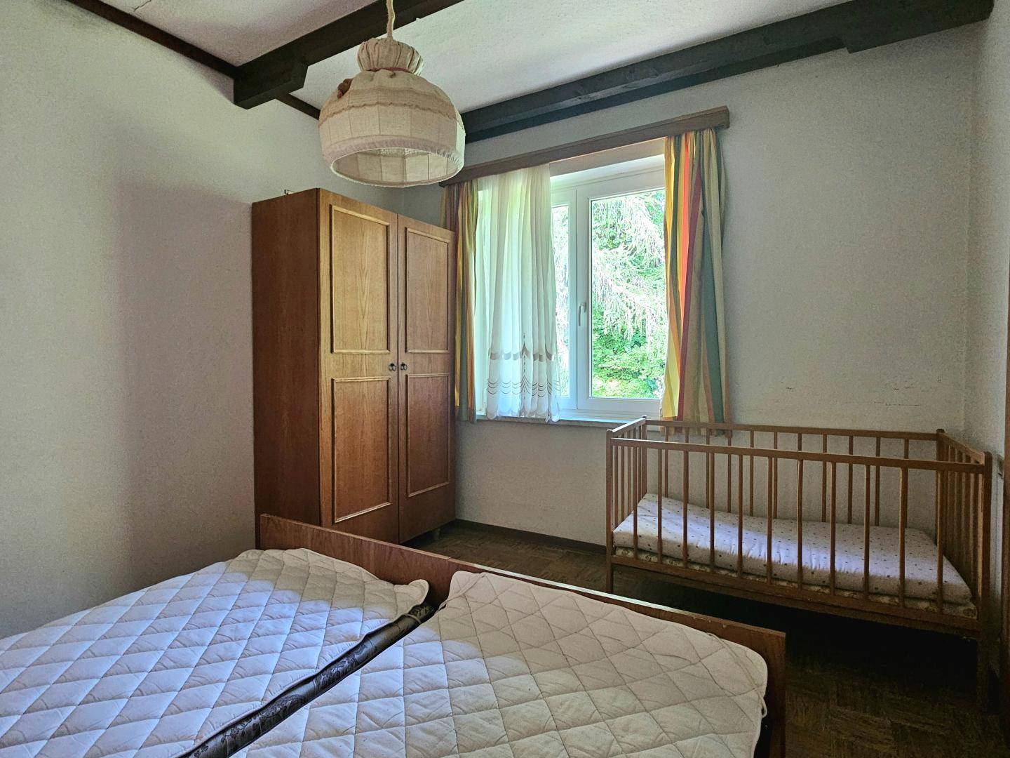 Schlafzimmer mit Holzschrank, Babybett und Fenster mit Blick auf die grüne Umgebung.