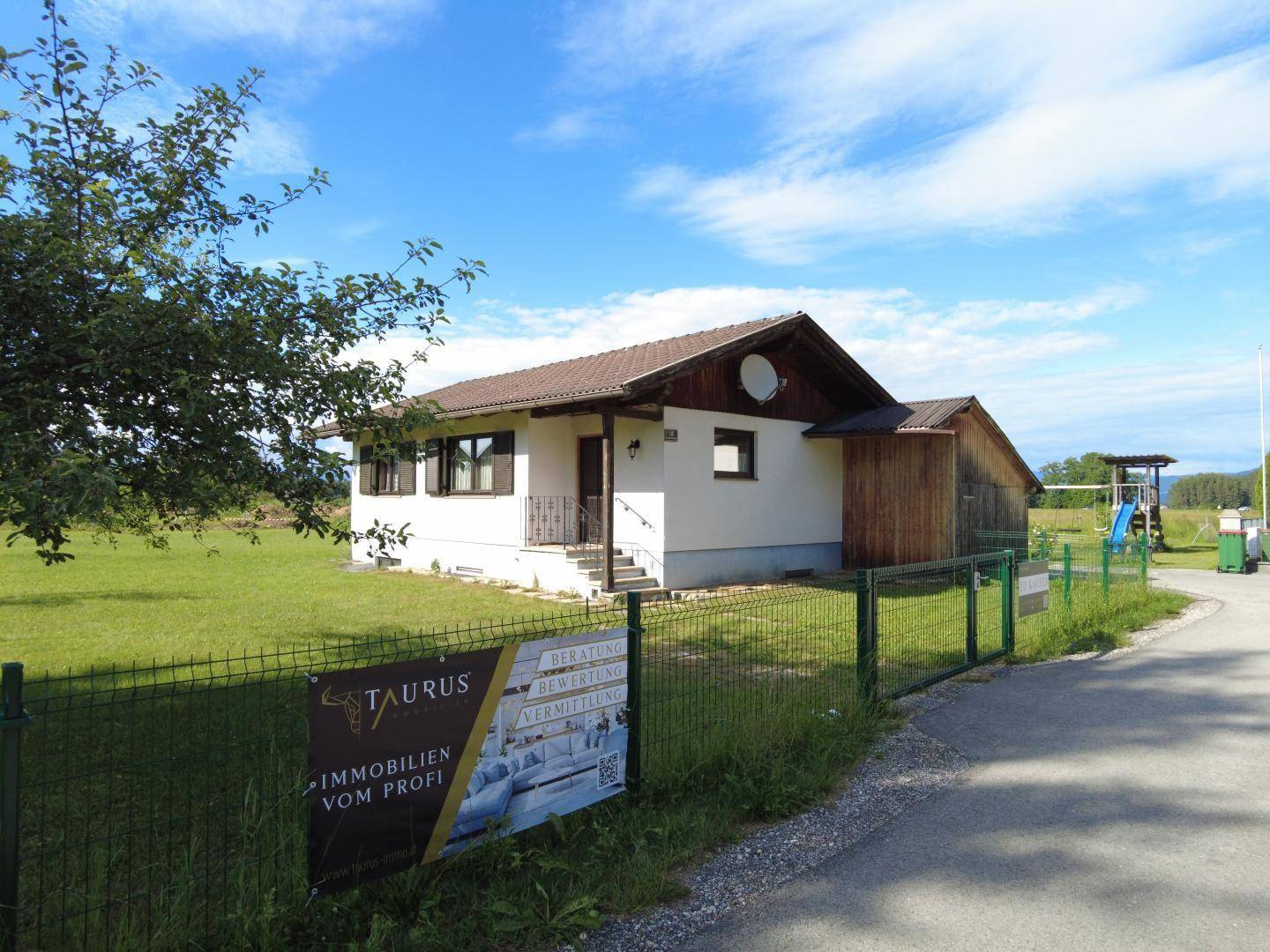 Einfamilienhaus mit Werbeschild am Zaun und Spielplatz im Hintergrund.