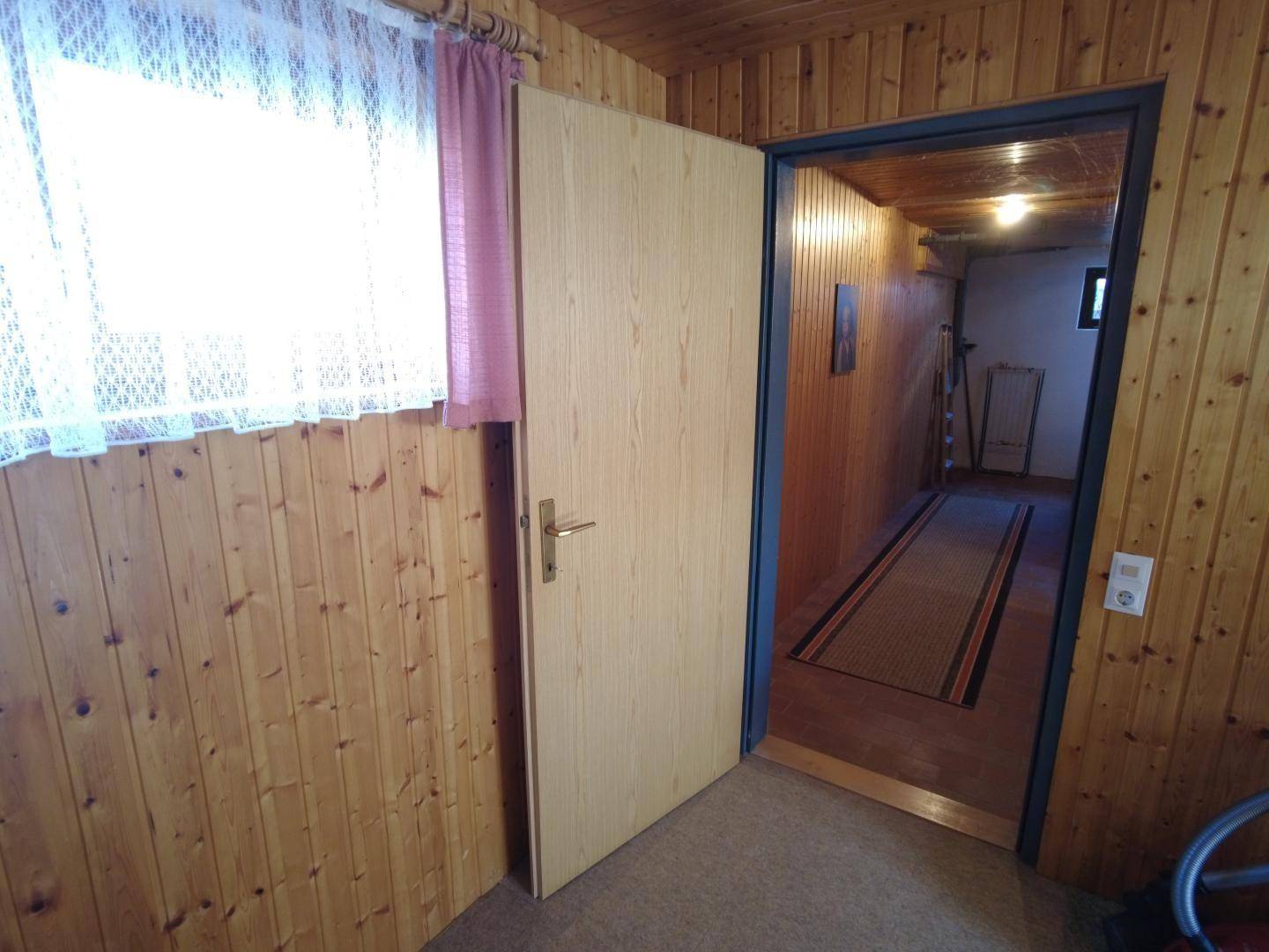 Tür in einem holzvertäfelten Raum mit Fenster und Vorhängen, die zu einem angrenzenden Flur führt.