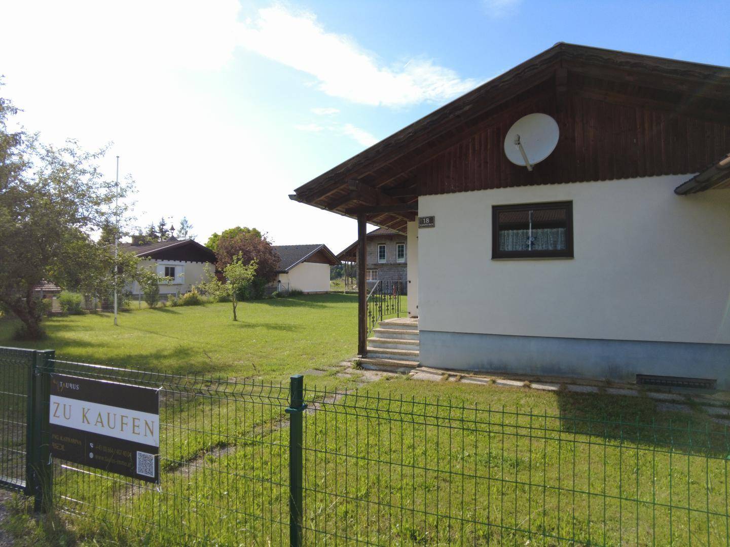 Einfamilienhaus mit "Zu Kaufen" Schild am Zaun und Blick auf die ländliche Umgebung.