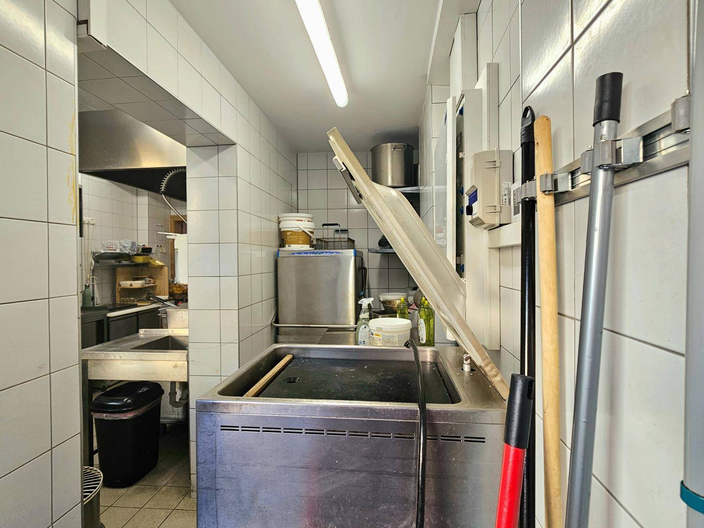 Schmaler Küchenbereich mit großem Spülbecken, Reinigungsutensilien und Zugang zu weiteren Arbeitsbereichen.
