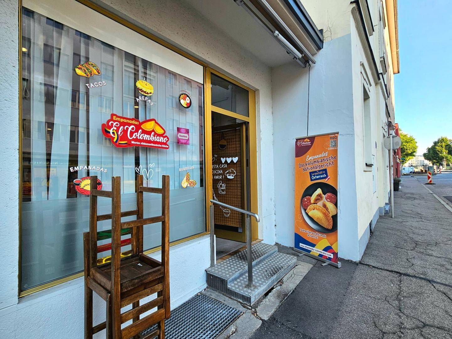Eingangsbereich des Restaurants mit Schaufenster, Werbebanner und einem Holzstuhl auf dem Bürgersteig.