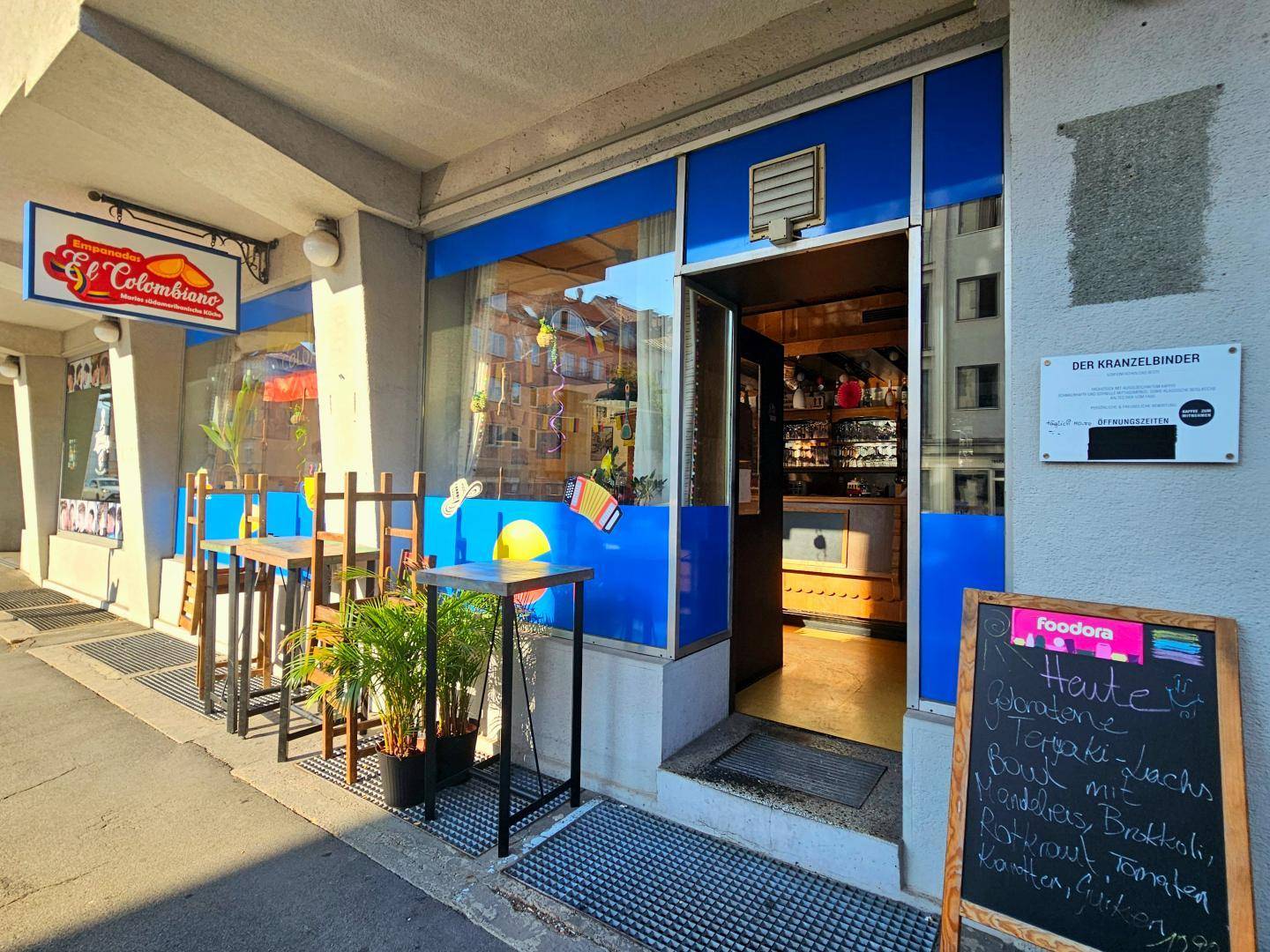 Außenansicht eines Restaurants mit blauer Fassade, Außentischen und einer Kreidetafel mit Tagesmenü.