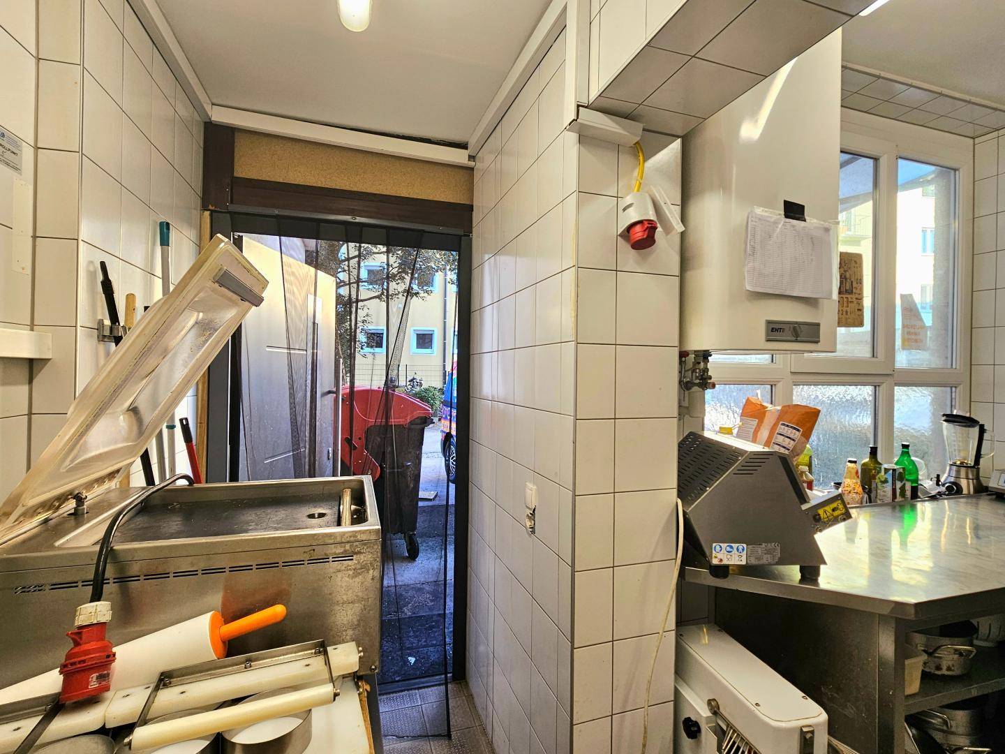 Blick aus einem schmalen Küchenbereich auf eine Ausgangstür, die ins Freie führt, mit Mülltonnen sichtbar.