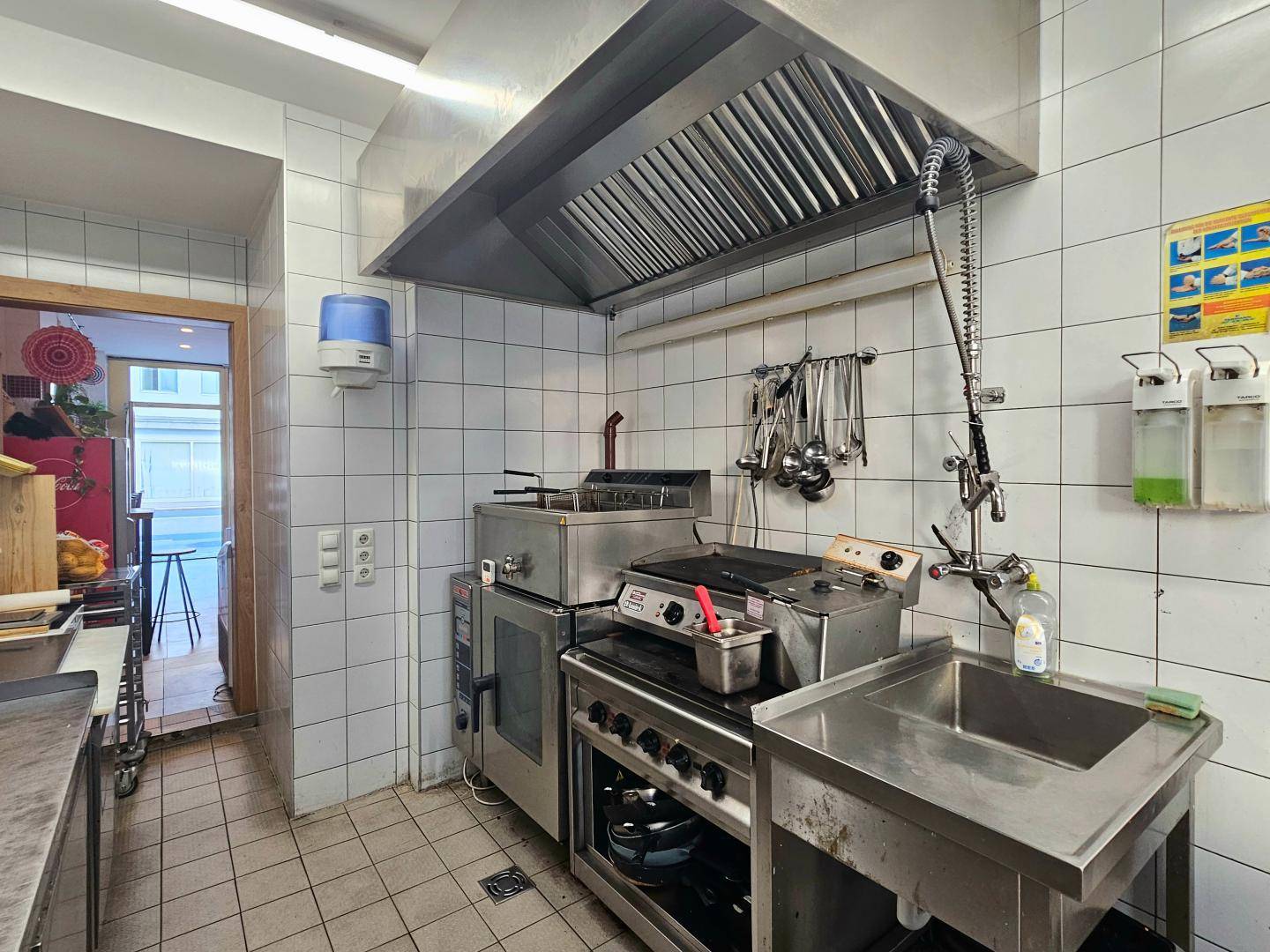 Industrielle Küche mit Fritteuse, Ofen, Spülbecken und einer großen Dunstabzugshaube über dem Kochbereich.