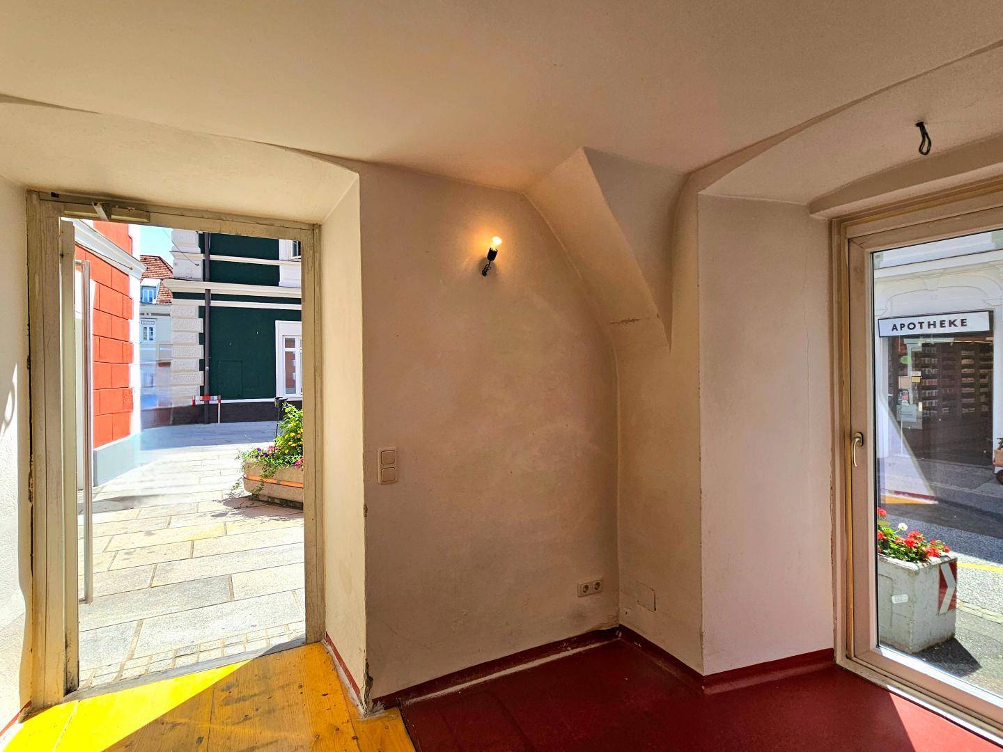 Heller Eingangsbereich mit Blick auf die Straße und einen Blumenkübel, roter Boden und weiße Wände.