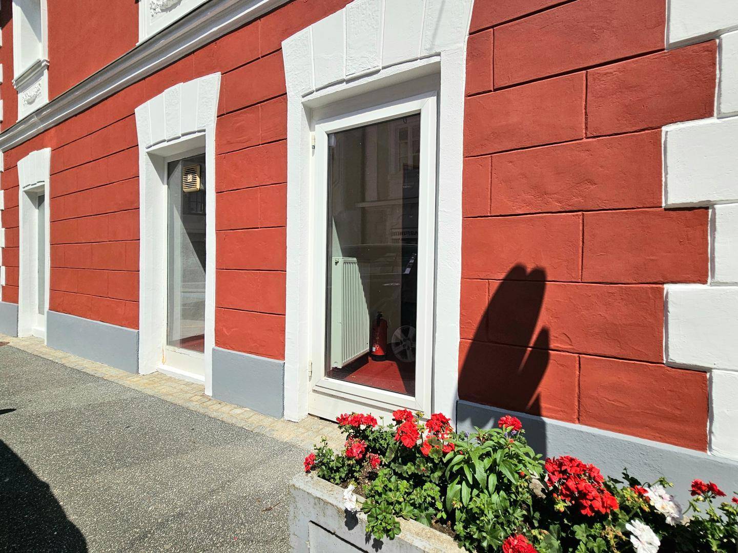 Nahaufnahme der roten Fassade mit weißen Fensterrahmen und einem Blumenkübel auf dem Bürgersteig.