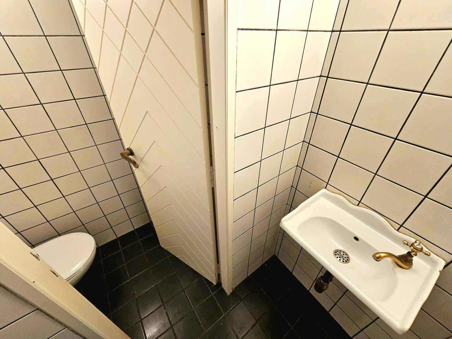Kleiner Toilettenraum mit weißen Fliesen an den Wänden und einem Waschbecken mit goldenem Wasserhahn.