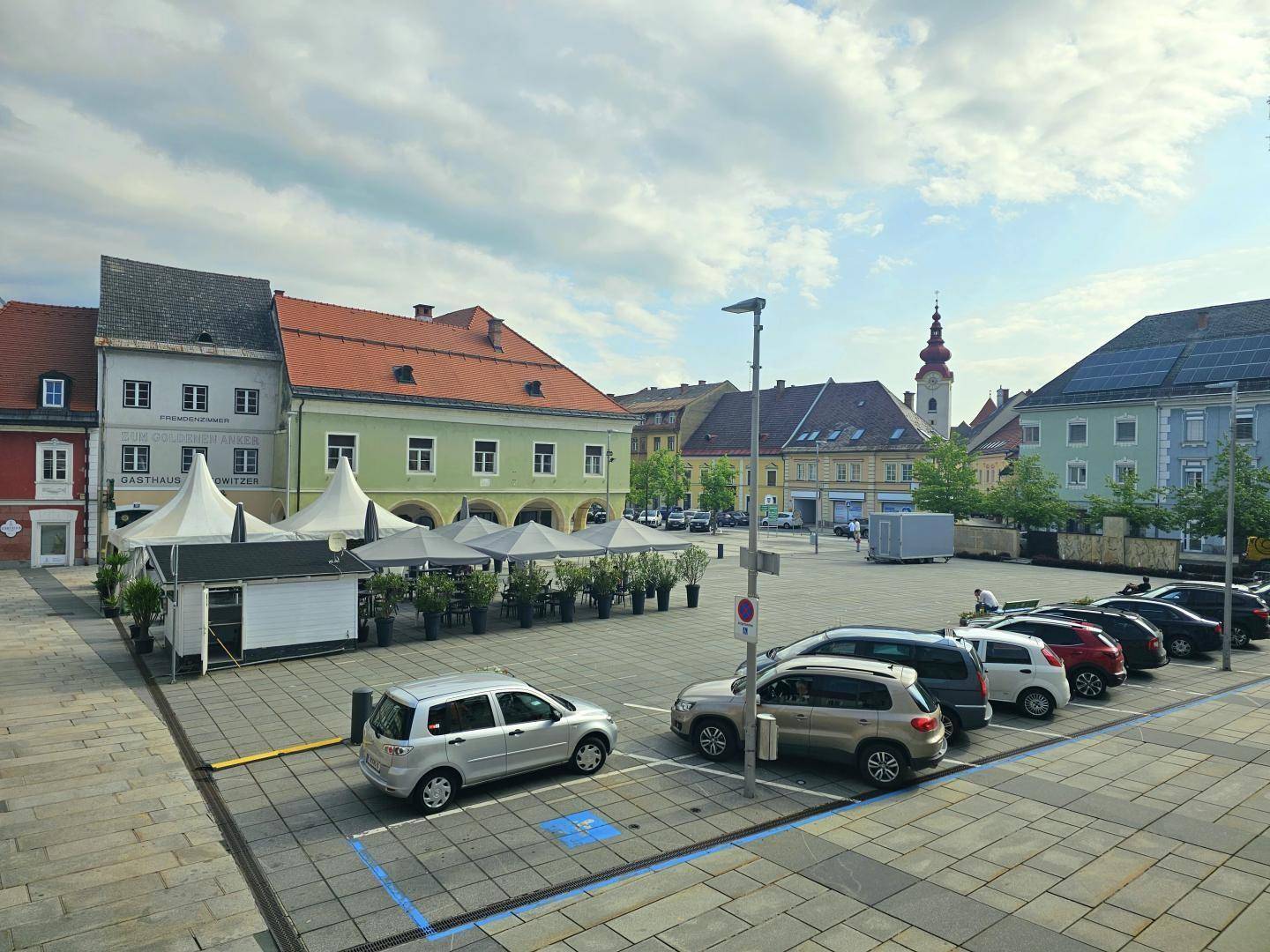 Blick auf einen belebten Stadtplatz mit parkenden Autos und umliegenden Gebäuden.