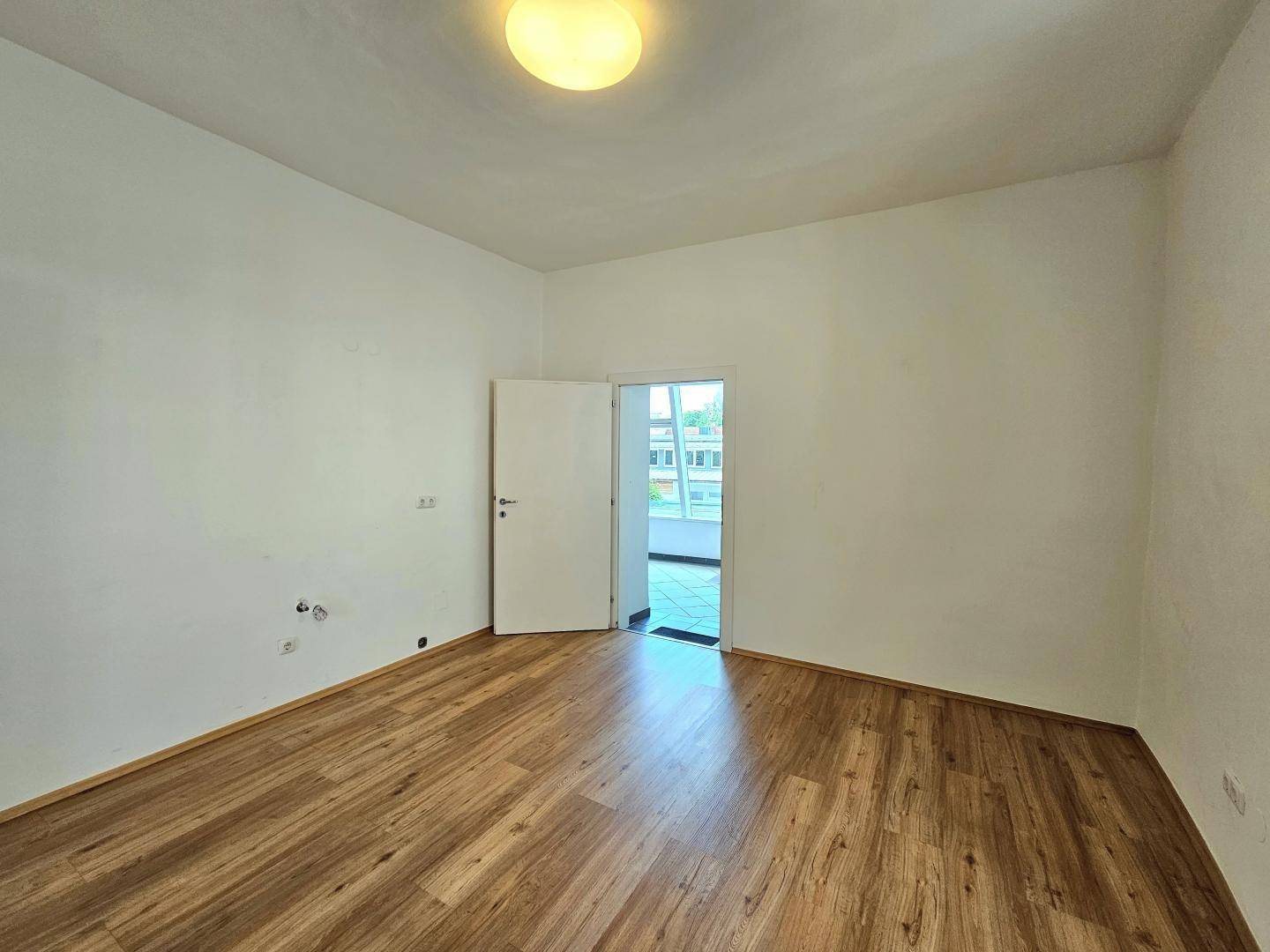 Leeres Zimmer mit Parkettboden und einer Tür, die zu einem helleren Bereich führt.