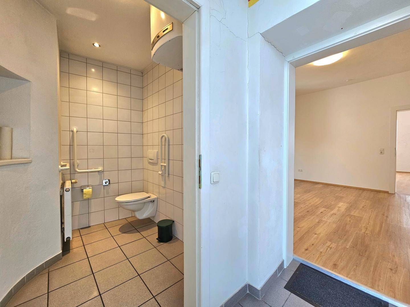 Barrierefreies WC mit Haltegriffen, Fliesenboden und Wand-WC.