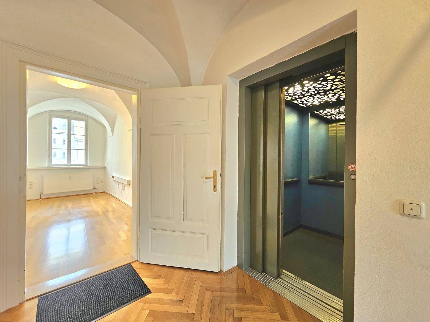 Heller Flur mit Aufzug und Blick in ein Zimmer mit Gewölbedecke und Fenster.