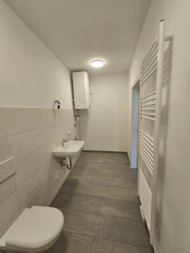 Langes Badezimmer mit Heizkörper, Wand-WC, Waschbecken und einem Warmwasserbereiter an der Wand.