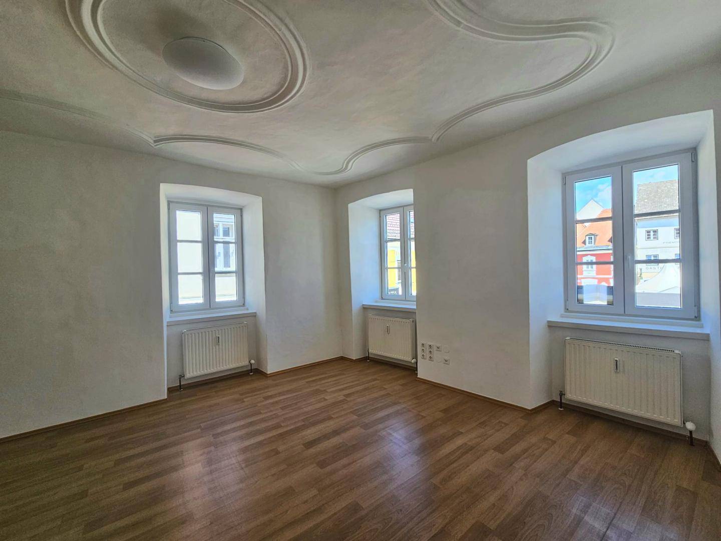 Helles Zimmer mit drei Fenstern, Parkettboden und Heizkörpern unter den Fenstern.