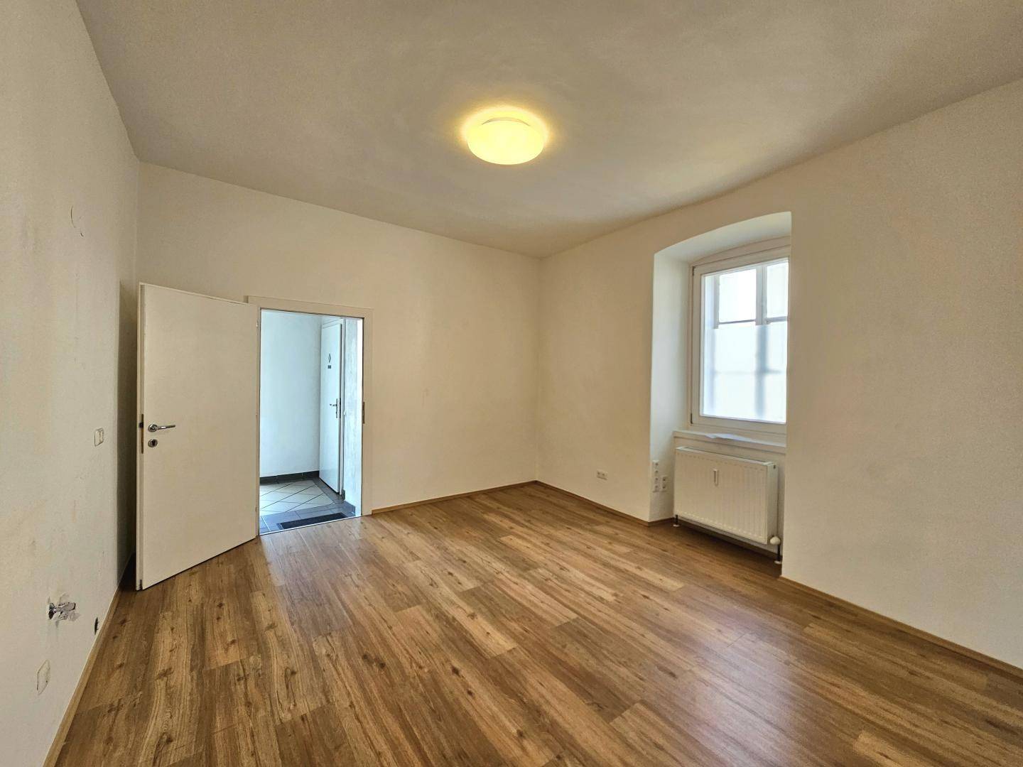 Helles Zimmer mit Parkettboden und einem Fenster, das viel Tageslicht hereinlässt.