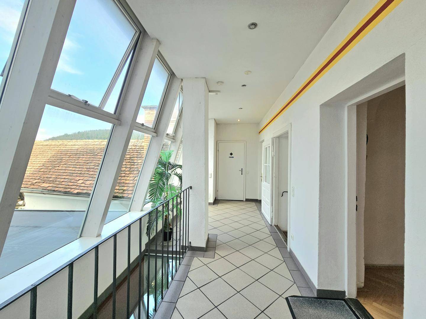 Langer, verglaster Balkon mit schrägen Fenstern und Zugang zu mehreren Türen im Innenbereich.
