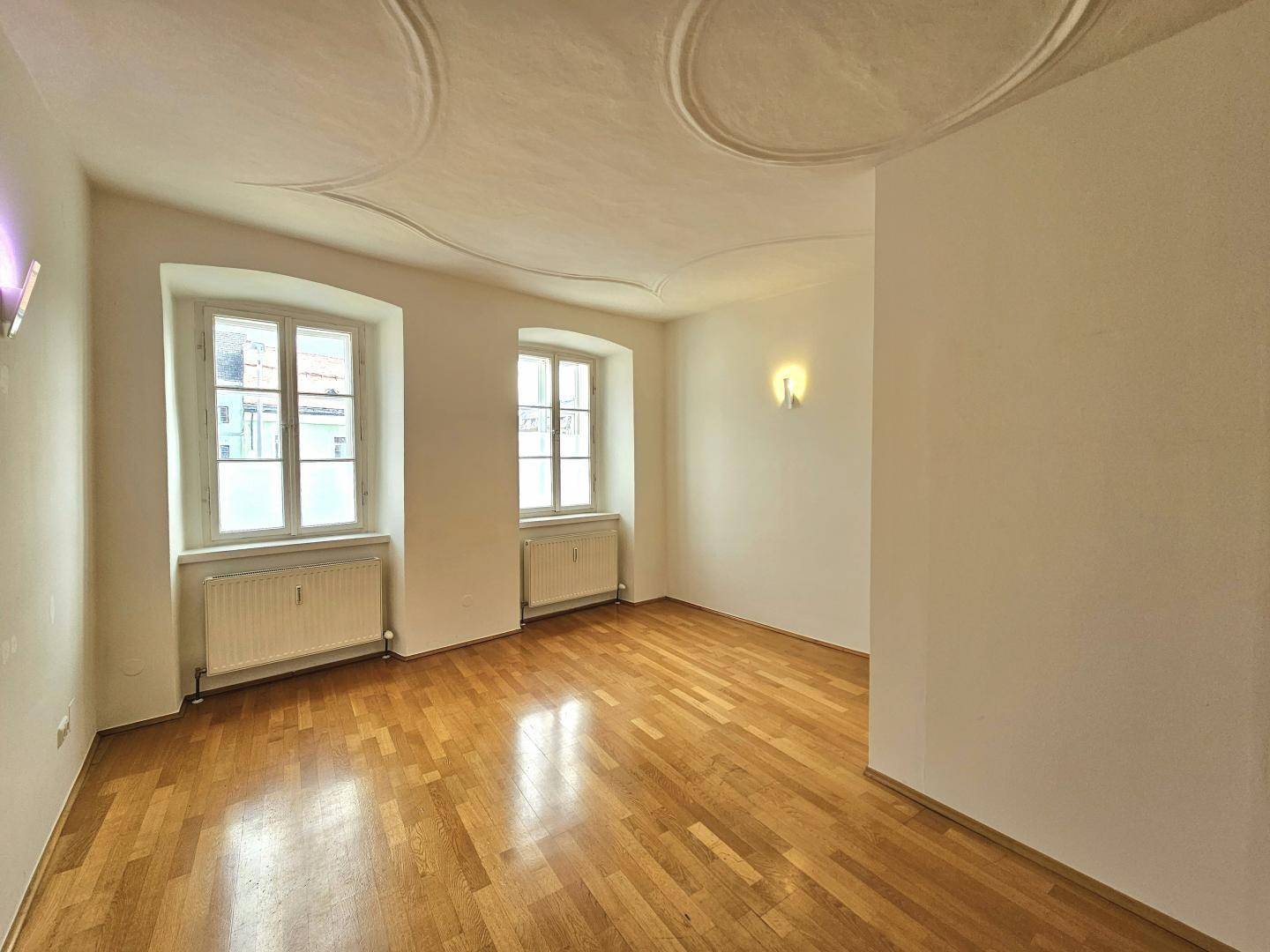 Helles Zimmer mit Parkettboden, zwei Fenstern und dekorativer Stuckdecke.
