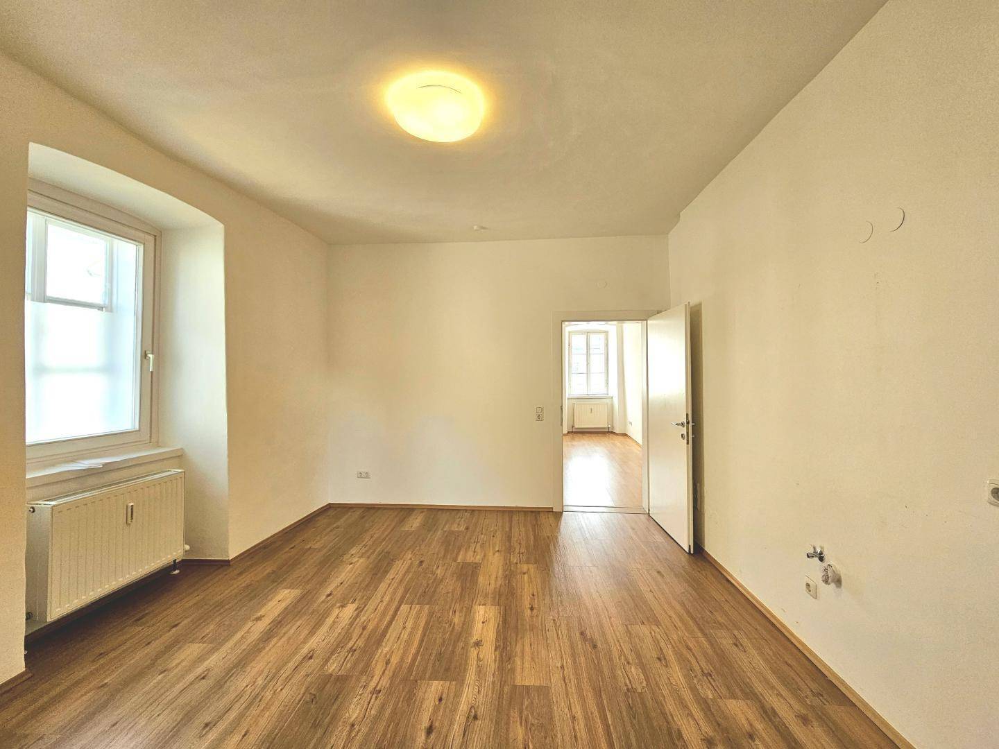 Helles Zimmer mit Parkettboden und einem Fenster, das viel Tageslicht hereinlässt.