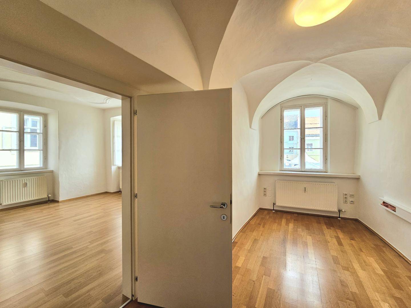 Zimmer mit Holzboden, Fenster und Gewölbedecke, verbunden durch eine offene Tür.