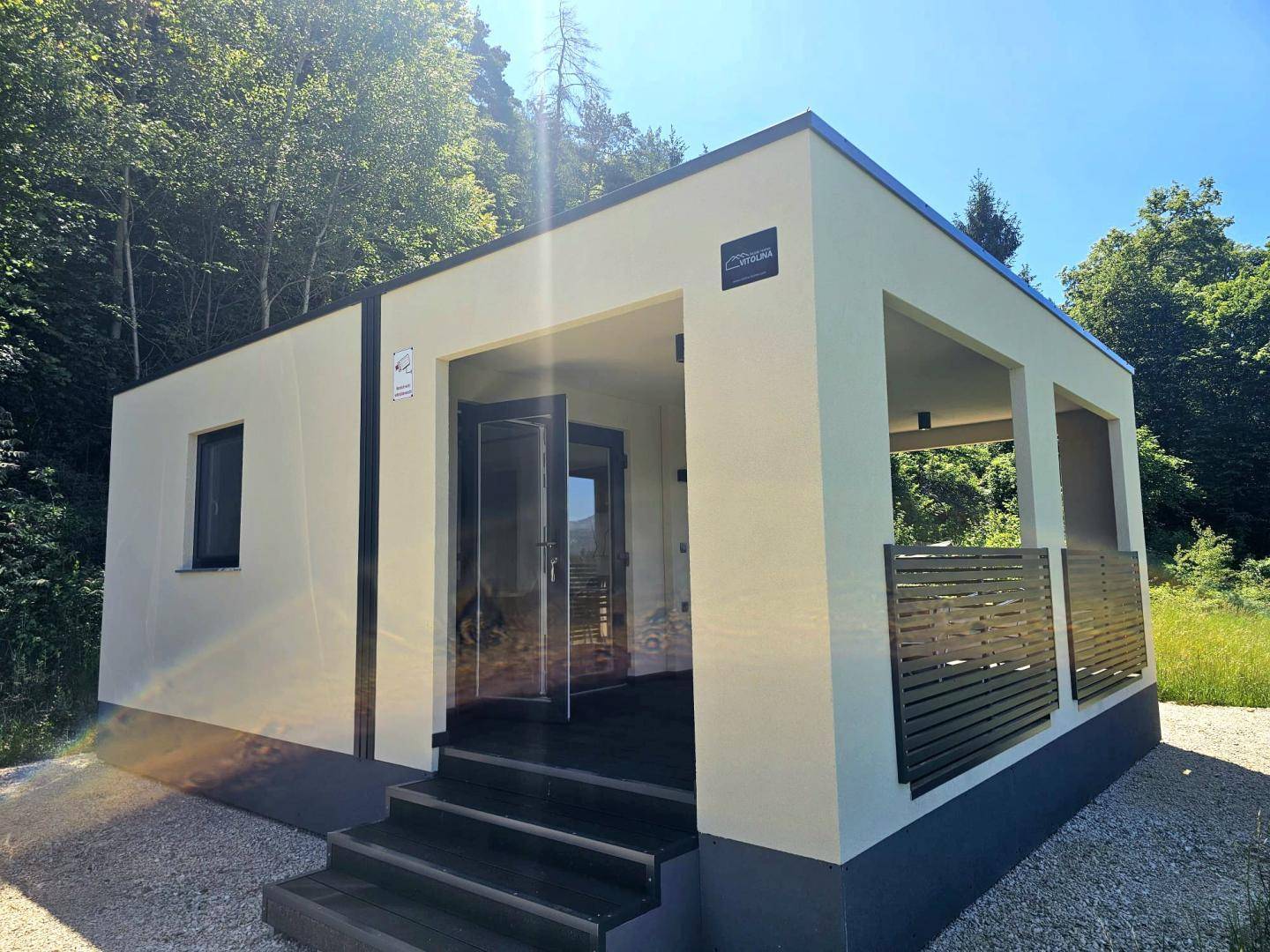 Eingangsbereich eines modernen Fertighauses mit dunkler Holzterrasse und umgebender Natur.