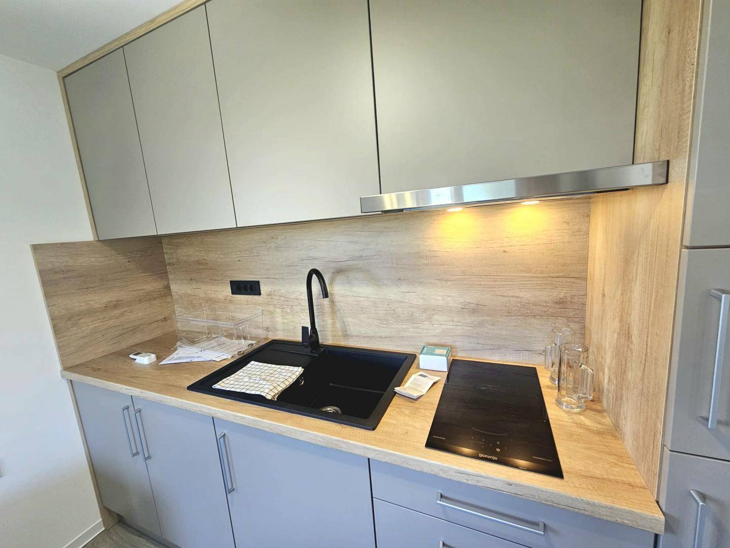 Moderne Küchenzeile mit Induktionskochfeld, Spüle und hellen Holz- und Grautönen.