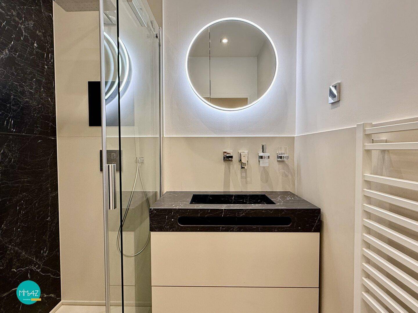 Elegantes Badezimmer mit begehbarer Dusche, dunklen Marmorfliesen und beleuchtetem Spiegel.