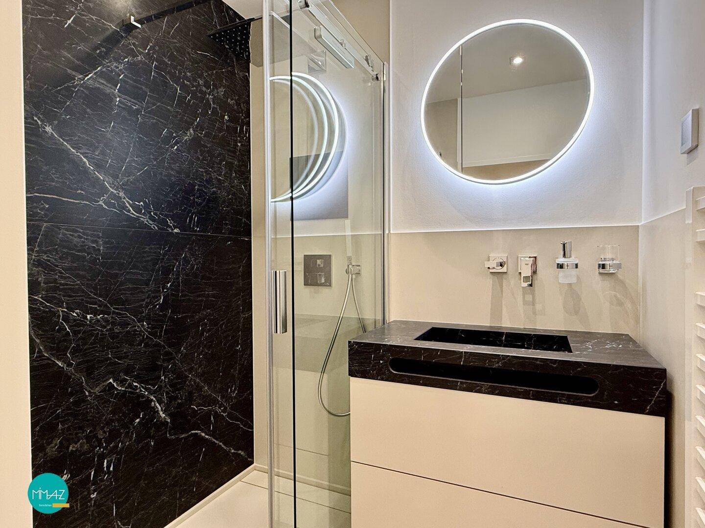 Elegantes Badezimmer mit begehbarer Dusche, dunklen Marmorfliesen und beleuchtetem Spiegel.