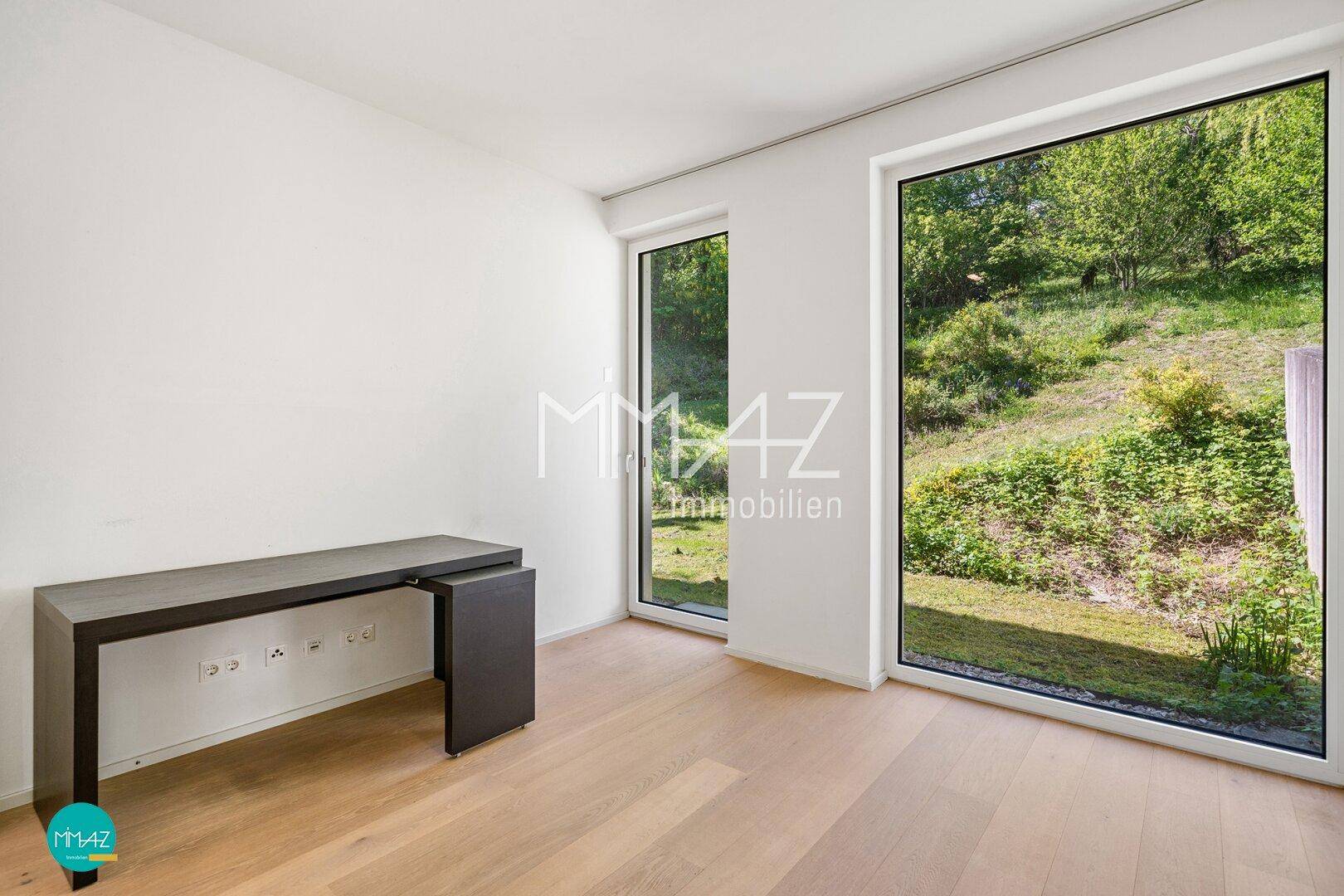 Helles Zimmer mit Holzboden und bodentiefen Fenstern, die einen Blick auf die grüne Umgebung bieten.