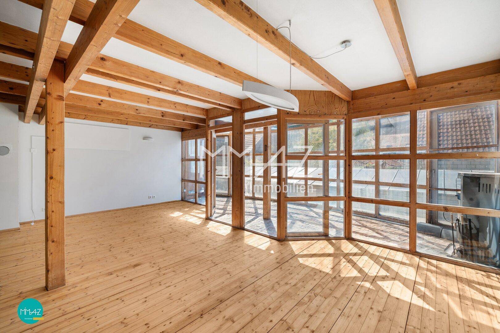 Heller, leerer Wohnbereich mit Holzbalken, Dielenboden und großen Fenstern zum Wintergarten.